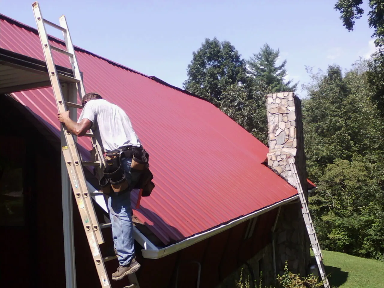 Roofer on a ladder beside a red metal roof and stone chimney — Soffit & Fascia Repair by Roofing Brothers in Entriken